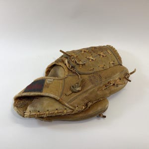 May include: A well-worn, tan leather baseball glove. The glove has visible stitching, laces, and a small patch with red and black text. The glove is designed to catch a baseball and is likely used for playing baseball.