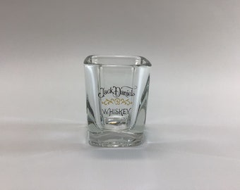 May include: A clear glass shot glass with a square base and a Jack Daniel's Whiskey logo printed on the front.