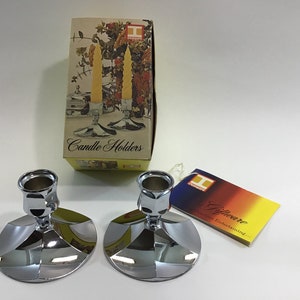 May include: Two silver candle holders with a round base and a fluted design. The candle holders are on a white surface. The box they came in is visible in the background. The box is yellow and has the words "Candle Holders" printed on it.
