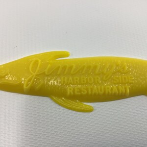 May include: Yellow plastic fish-shaped sign with the text "Jimmy's Harbor Side Restaurant" embossed on it.