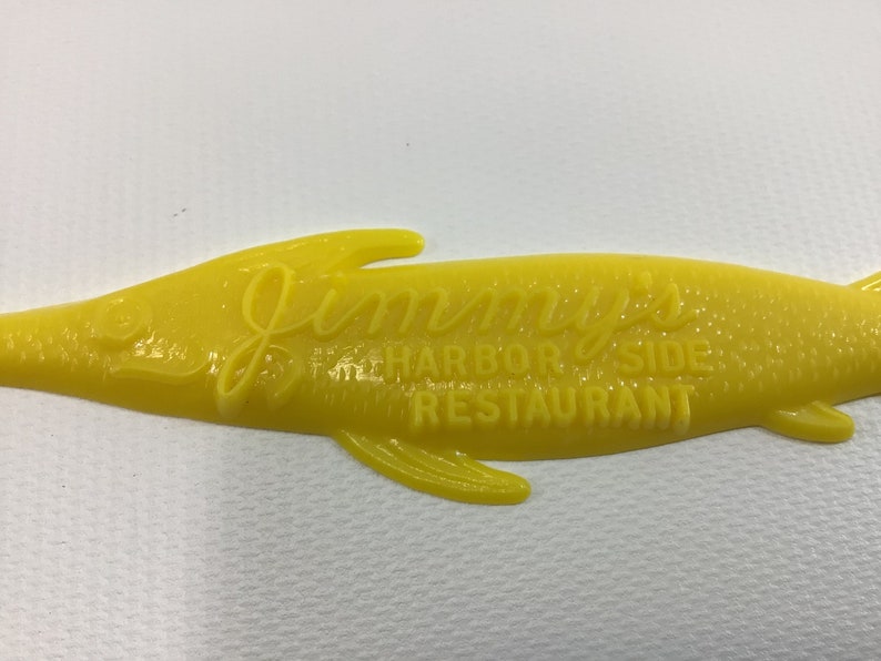 May include: Yellow plastic fish-shaped sign with the text "Jimmy's Harbor Side Restaurant" embossed on it.