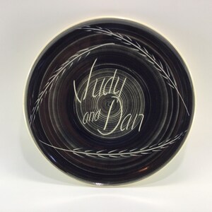 May include: A dark blue plate with a spiral design and the names "Judy and Dan" in white script. The plate has a white rim and a decorative white leaf design around the edge. A decorative plate.
