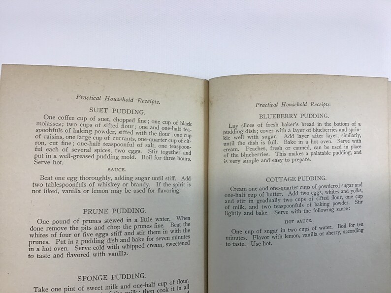 May include: An open vintage cookbook with recipes for Suet Pudding, Blueberry Pudding, Cottage Pudding, Prune Pudding, and Sponge Pudding. The book has a yellowed, cream-colored paper cover with black text.