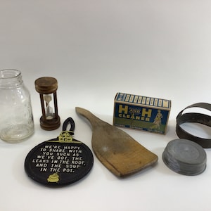 May include: A collection of vintage household items including a glass jar, an hourglass, a wooden spatula, a cast iron trivet with a humorous saying, a box of "H & H Cleaner", a metal ring mold, and a metal lid.