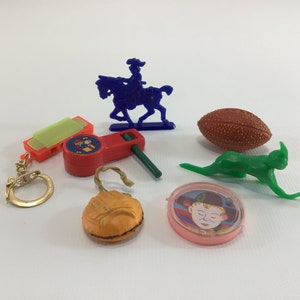 May include: A collection of vintage plastic toys, including a blue plastic toy horse and rider, a green plastic toy rabbit, a red plastic toy whistle, a brown plastic toy football, a yellow plastic toy harmonica, a gold key chain, a pink plastic toy with a face, and an orange plastic toy shaped like a baseball mitt.