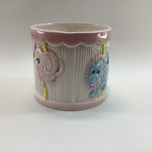 May include: A pink and white ceramic planter with a carousel design featuring a pink elephant, a blue lion, and yellow poles.