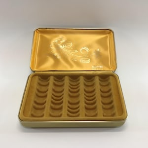 May include: Open gold-toned jewelry box with a velvet interior. The box has a rectangular shape with a hinged lid. The inside of the lid has a gold satin lining with a decorative design. The base has multiple curved slots.