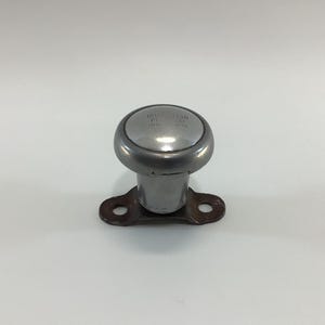 May include: A vintage, silver-toned steering wheel spinner knob. The knob has a rounded top with embossed text and a cylindrical base attached to a brown, metal mounting plate with two holes. The object is set against a white background.