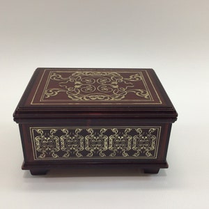 May include: A wooden music box with a dark brown finish and gold inlay. The top of the box features a decorative design with intricate swirls and scrolls. The sides of the box have a repeating pattern of smaller swirls.