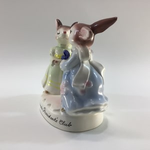 May include: A porcelain figurine of two bunnies dressed in pastel-colored dresses. The bunny on the right is wearing a blue dress with a white sash and a pink flower on her dress. The bunny on the left is wearing a green dress with a yellow sash. The base of the figurine is white and has the words "Presidents' Club" written on it.