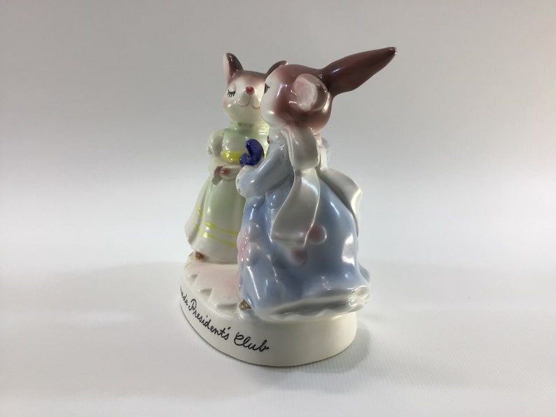 May include: A porcelain figurine of two bunnies dressed in pastel-colored dresses. The bunny on the right is wearing a blue dress with a white sash and a pink flower on her dress. The bunny on the left is wearing a green dress with a yellow sash. The base of the figurine is white and has the words "Presidents' Club" written on it.
