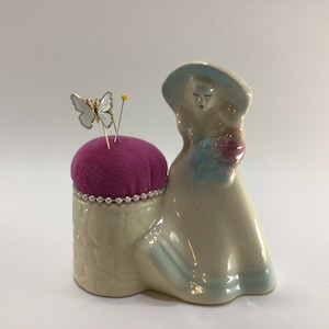May include: A ceramic pincushion shaped like a woman in a white dress and blue hat. The woman is holding a bouquet of flowers. The pincushion has a pink velvet top and a white base with a pearl trim. A white butterfly is pinned to the top.