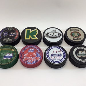 May include: Eight round hockey pucks with various designs. The pucks feature the Kalamazoo K-Wings logo, a green shamrock, a heart design, a 25th anniversary logo, a Hershey's chocolate logo, and a Michigan K-Wings logo.