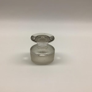 May include: A clear glass bottle stopper with a wide, flat, circular top and a tapered neck. The base of the stopper is cylindrical with a frosted, translucent finish, designed to fit securely into the opening of a glass bottle or decanter.