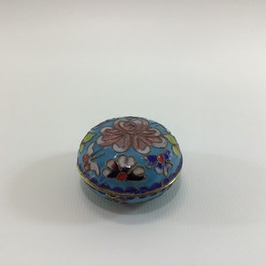 May include: A small, round, blue and white enamel trinket box with a floral design. The box is decorated with pink, green, yellow, and blue flowers.