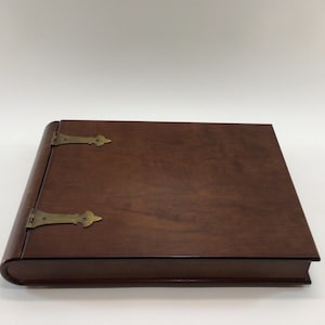May include: A closed, rectangular wooden box with a dark brown finish. The box has rounded edges and is secured with two brass-colored hinges. The overall design suggests a vintage or antique style, suitable for storage or display.