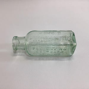 May include: A clear, rectangular glass bottle with rounded edges, embossed with text. The bottle has a short neck and a slightly greenish tint. The text reads "Household Extract" and "For Making Root Beer".
