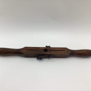 May include: A vintage wooden drawknife with a dark brown finish. The tool has two rounded handles and a central blade housing with metal hardware. It is designed for shaping wood.