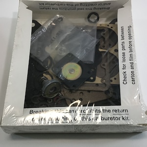 May include: A white box with a clear plastic window showing a carburetor kit. The kit includes a variety of parts, including gaskets, springs, and a metal float. The box has text that reads "Breaking this seal prohibits the return and/or crediting of this carburetor kit." and "Check for loose parts between carton and film before opening."