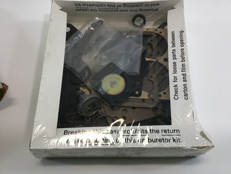 May include: A white box with a clear plastic window showing a carburetor kit. The kit includes a variety of parts, including gaskets, springs, and a metal float. The box has text that reads "Breaking this seal prohibits the return and/or crediting of this carburetor kit." and "Check for loose parts between carton and film before opening."