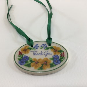 May include: Oval ceramic tag with a green ribbon. The tag is decorated with yellow and blue flowers and the words "Thank You".