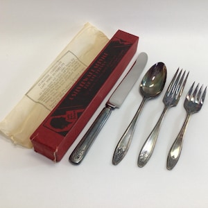 May include: A vintage silverware set with a knife, spoon, and two forks. The set includes a red box with text and a paper with text. The silverware has a floral design on the handles.