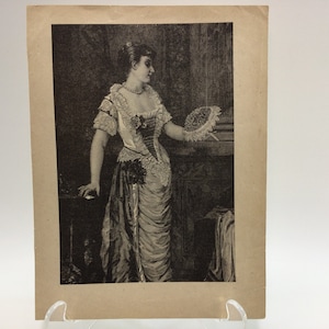 May include: A black and white print of a woman in a long, flowing dress holding a fan. She is standing in a room with a dark background.