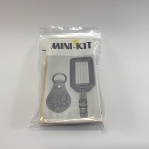 May include: A white plastic bag containing a leather craft kit with the text "MINI-KIT" printed on the front. The kit includes a leather key fob with a butterfly design and a leather luggage tag with a blank space for writing.