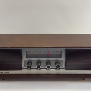 Puede incluir: Radio Ross vintage con una carcasa de chapa de madera oscura. La radio tiene una cara plateada con un dial de sintonización, indicadores de emisoras y mandos de control. Las rejillas de los altavoces son de color marrón oscuro.