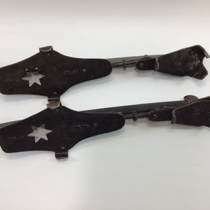 Könnte beinhalten: Ein Paar antike Eislaufschuhe aus Metall mit einem sternförmigen Ausschnitt an der Spitze. Die Schlittschuhe sind verstellbar und haben eine schwarze Oberfläche.