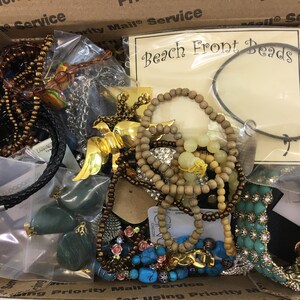 May include: A box filled with various necklaces, bracelets, and other jewelry pieces. The jewelry is made of different materials, including wood, metal, and beads. The box is labeled "Beach Front Beads".