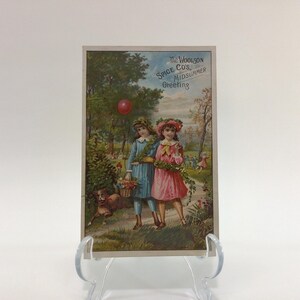 May include: Vintage postcard featuring two children in a woodland setting. The girl on the left is wearing a blue dress and holding a basket. The girl on the right is wearing a pink dress and holding a wreath. The text on the postcard reads "The Woolson Spice Co.'s Midsummer Greeting."