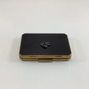 May include: A vintage compact with a black enamel top and gold-toned metal frame. A small, decorative cluster of rhinestones adorns the center of the compact. The rectangular shape and clasp closure suggest a classic design, suitable for cosmetics.