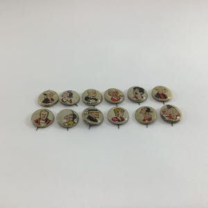 May include: A collection of twelve vintage pinback buttons with cartoon character illustrations. The buttons feature various characters in a range of colors, set against a light gray background. The buttons are round and appear to be made of metal.