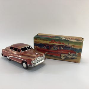 May include: A vintage toy car in a reddish-brown hue, with a chrome grill and accents. The car is next to its original box, which features an illustration of a red sedan. The box reads "Firestone Sedan".