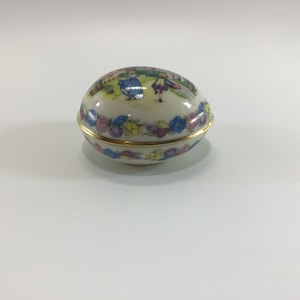 May include: A white porcelain egg-shaped trinket box with a gold trim and a floral design. The box features a painted scene of children playing in a garden.