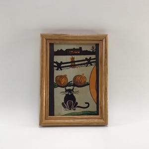 May include: A framed Halloween-themed print featuring a black cat, pumpkins, and a fence. The artwork has a rustic style with a light brown wooden frame. The background shows a house and a large orange moon.