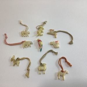 May include: A collection of small, off-white animal charms with attached cords. The charms include a bear, a kangaroo, a deer, a sheep, a parrot, a donkey, and a camel. The parrot has red, blue, and yellow coloring. The cords are various colors.