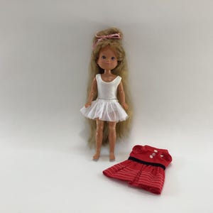 May include: A vintage doll with long blonde hair, wearing a white leotard and tutu. A red dress with white stripes and black trim is next to the doll. The doll has a pink bow in its hair.