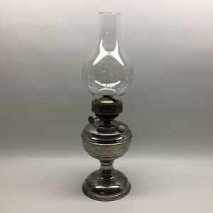 May include: A silver-toned metal oil lamp featuring a rounded base and a pedestal stand. The lamp includes a dark metal burner assembly with a circular adjustment knob and a clear glass chimney globe with a subtle etched floral pattern design.