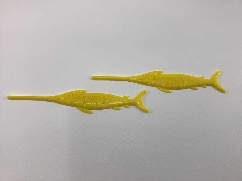 May include: Two yellow plastic fish-shaped cocktail picks with the text "Jimmy's Seafood Restaurant" printed on them.