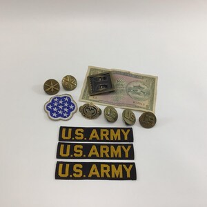 May include: A collection of U.S. Army memorabilia, including patches, buttons, and a bill. The patches are black with gold lettering. The buttons are various sizes and shapes, with a mix of gold and bronze tones. A blue and white patch with stars is also present.