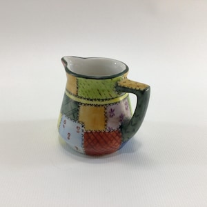 May include: A small, patchwork-patterned ceramic pitcher with a handle. The pitcher features a variety of colors, including green, yellow, blue, and red, arranged in a quilt-like design. The handle is dark green, and the interior is white.