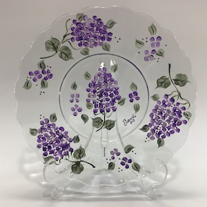 May include: A clear glass plate with a scalloped edge, hand-painted with purple and green lilac flowers. The plate is signed "Diane '93" in the center.