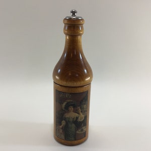 May include: A wooden bottle-shaped salt and pepper shaker with a metal top. The bottle is decorated with a vintage Coca-Cola advertisement featuring a woman in a hat.