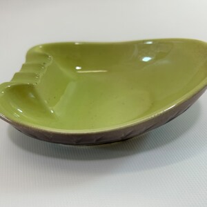 May include: A green ceramic ashtray with a brown rim and a leaf design. The ashtray has four indentations for cigarettes.