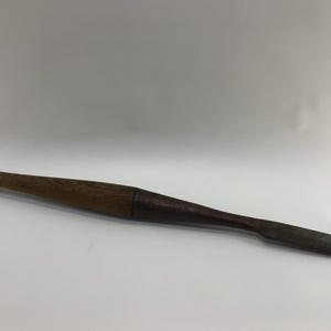 May include: A long, wooden tool with a rounded handle and a tapered, flat end. The wood has a dark brown color, with variations in tone along its length. The tool appears to be handmade and is set against a white background.
