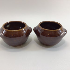 May include: Two brown ceramic bowls with handles. The bowls are round and have a glossy finish.