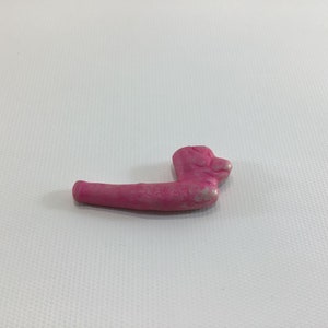 May include: A pink ceramic pipe shaped like a dog's head. The pipe is about 5 inches long and has a textured surface.
