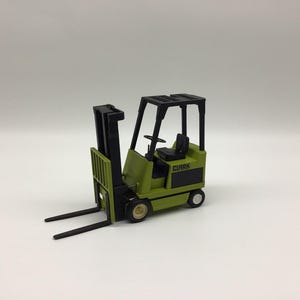 May include: A miniature olive green forklift model featuring a black mast, black overhead guard, and black seat. The side of the vehicle displays the brand name CLARK in black lettering. It has four small wheels and two long, thin black forks.